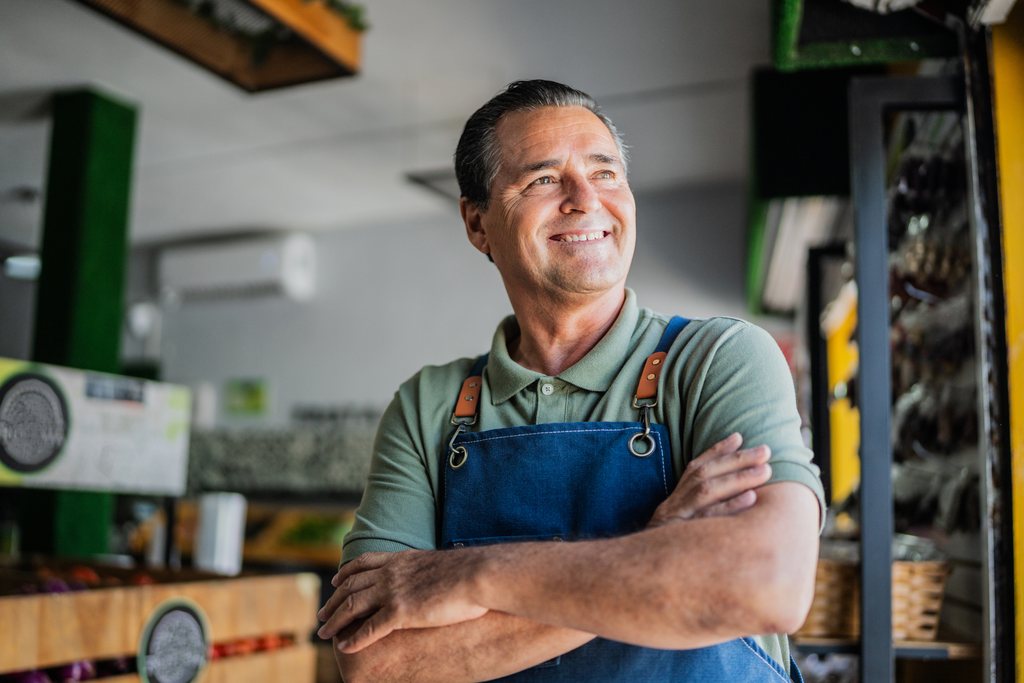 A shop owner in a denim apron, arms folded, smiling toward the door of his store